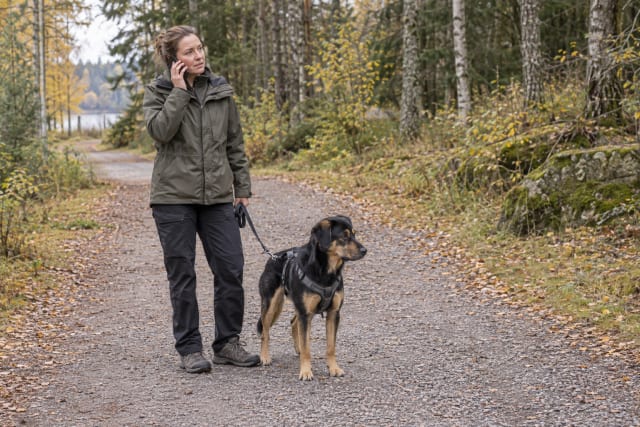 Hundägare talar i telefon med en kopplad hund bredvid sig på en öppen stig.